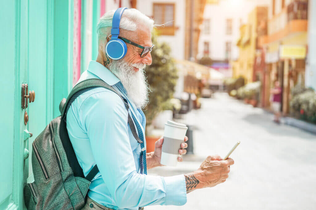 Trendy senior man using music smartphone app and drinking coffee in downtown center - Mature fashion male having fun with new trends technology.