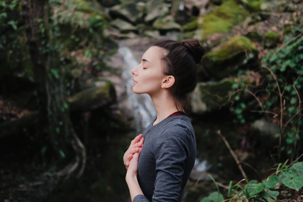 Young woman practicing breathing yoga pranayama outdoors in moss forest on background of waterfall.